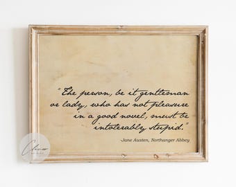 May include: Framed print featuring a quote from Jane Austen's "Northanger Abbey" in elegant black script. The quote is set against a vintage-style parchment background, encased in a light brown wooden frame. The quote reads: "The person, be it gentleman or lady, who has not pleasure in a good novel, must be intolerably stupid."