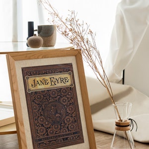 May include: A framed print of the book cover for "Jane Eyre" with a decorative floral design and the text "Household Edition". The print is on a table with a vase of dried flowers and a ceramic mug.