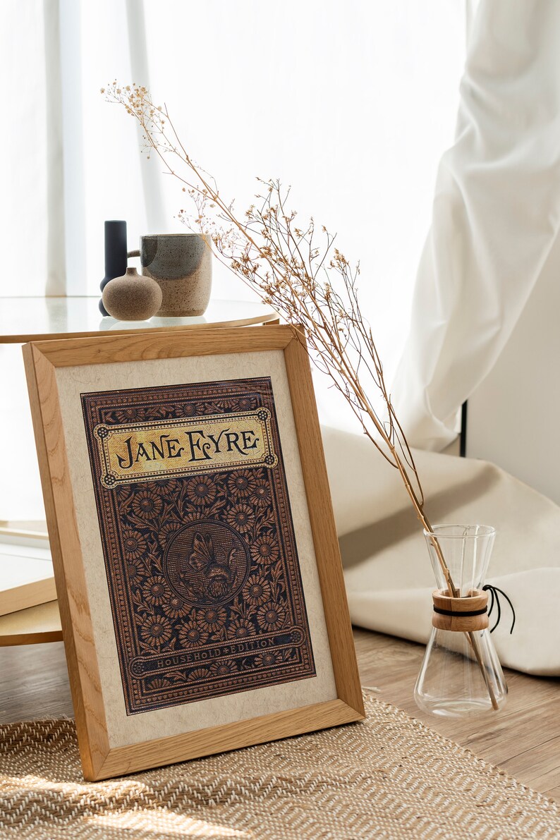 May include: A framed print of the book cover for "Jane Eyre" with a decorative floral design and the text "Household Edition". The print is on a table with a vase of dried flowers and a ceramic mug.