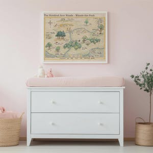 May include: A white dresser with two drawers, topped with a pink changing pad. Above the dresser hangs a framed map of The Hundred Acre Woods. A wicker basket and a small tree in a woven pot complete the nursery decor.
