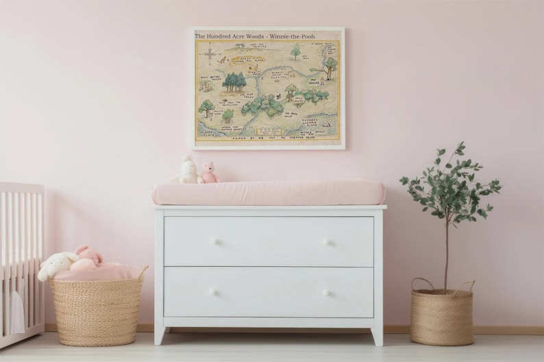 May include: A white dresser with two drawers, topped with a pink changing pad. Above the dresser hangs a framed map of The Hundred Acre Woods. A wicker basket and a small tree in a woven pot complete the nursery decor.