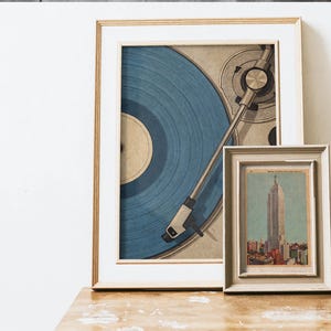 May include: Two framed art prints. The larger print features a blue vinyl record on a turntable. The smaller print depicts a cityscape with a tall building. Both prints are in neutral-toned frames, resting on a wooden surface.