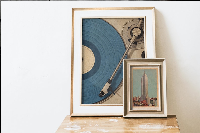 May include: Two framed art prints. The larger print features a blue vinyl record on a turntable. The smaller print depicts a cityscape with a tall building. Both prints are in neutral-toned frames, resting on a wooden surface.