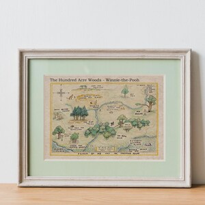 May include: Framed print of a map titled "The Hundred Acre Woods - Winnie-the-Pooh." The map features illustrations of trees, rivers, and landmarks, with text labels. The frame is white with a light green mat.