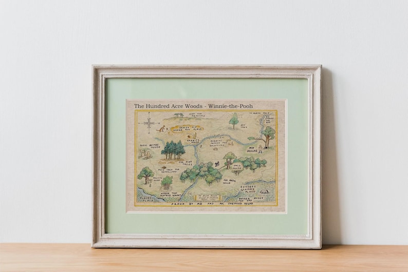 May include: Framed print of a map titled "The Hundred Acre Woods - Winnie-the-Pooh." The map features illustrations of trees, rivers, and landmarks, with text labels. The frame is white with a light green mat.