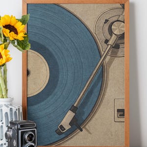 May include: A framed print of a vintage record player with a blue vinyl record. The artwork has a retro aesthetic, with a wooden frame and a neutral background. A vase of sunflowers and a vintage camera are in the foreground.