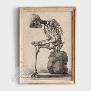 May include: Framed vintage print of a human skeleton in a seated position, resting on a stone. The black and white anatomical illustration is set against a beige background, and encased in a gold-toned wooden frame. The skeleton's posture suggests contemplation.