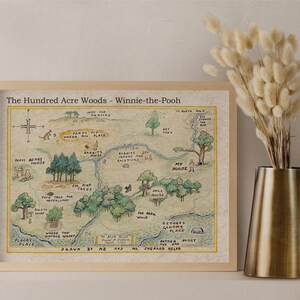 May include: Framed print of The Hundred Acre Woods map from Winnie-the-Pooh, with a light wood frame. The map is hand-drawn with watercolor-style illustrations and text. A brass pitcher with dried flowers is to the right.