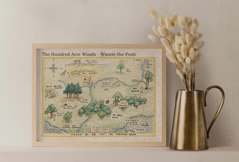 May include: Framed print of The Hundred Acre Woods map from Winnie-the-Pooh, with a light wood frame. The map is hand-drawn with watercolor-style illustrations and text. A brass pitcher with dried flowers is to the right.