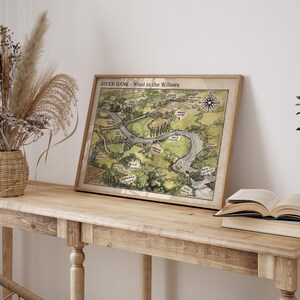 May include: A framed map titled "River Bank - Wind in the Willows" with a compass rose, depicting a river and surrounding landscape in shades of green and brown. The map is displayed on a light-colored wooden table, next to a vase of dried flowers and an open book.