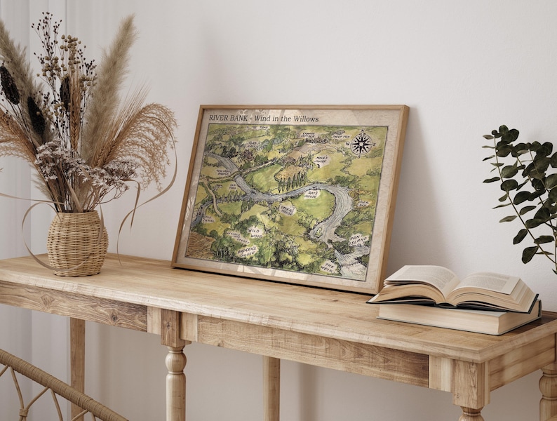 May include: A framed map titled "River Bank - Wind in the Willows" with a compass rose, depicting a river and surrounding landscape in shades of green and brown. The map is displayed on a light-colored wooden table, next to a vase of dried flowers and an open book.