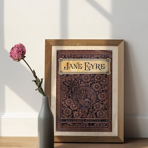 May include: A framed print of the book cover for "Jane Eyre" with a decorative floral design in gold and brown on a black background. The title "Jane Eyre" is in gold lettering. The text "Household Edition" is at the bottom of the cover.