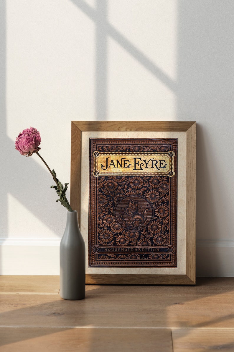 May include: A framed print of the book cover for "Jane Eyre" with a decorative floral design in gold and brown on a black background. The title "Jane Eyre" is in gold lettering. The text "Household Edition" is at the bottom of the cover.