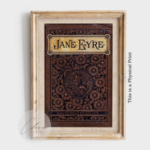May include: Framed print of a vintage book cover. The cover is dark brown with a floral pattern and the title "JANE EYRE" in gold lettering. The words "HOUSEHOLD EDITION" are at the bottom. The frame is light brown.