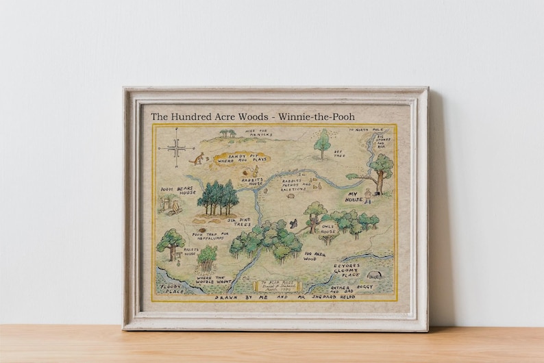 May include: Framed map of The Hundred Acre Woods from Winnie-the-Pooh, featuring a hand-drawn style with whimsical illustrations of locations like Rabbit's House and Pooh's house. The map is in a white frame, resting on a wooden surface.