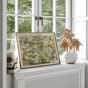May include: A framed map titled "RIVER BANK - Wind in the Willows" depicting a river and surrounding landscape. The map is in a wooden frame and rests on a white windowsill. A white vase with dried plants sits nearby.