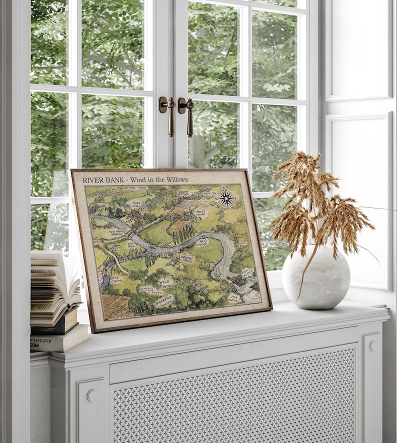 May include: A framed map titled "RIVER BANK - Wind in the Willows" depicting a river and surrounding landscape. The map is in a wooden frame and rests on a white windowsill. A white vase with dried plants sits nearby.