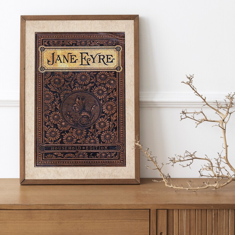 May include: A framed print of the book cover for "Jane Eyre: Household Edition". The cover features a dark brown background with a gold floral pattern and the title "Jane Eyre" in gold lettering.