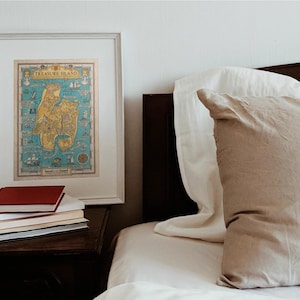 May include: A framed map of Treasure Island with a blue background and yellow and brown land masses. The map is hanging on a white wall above a nightstand with a stack of books. A bed with white and beige pillows is in the background.