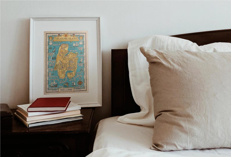 May include: A framed map of Treasure Island with a blue background and yellow and brown land masses. The map is hanging on a white wall above a nightstand with a stack of books. A bed with white and beige pillows is in the background.