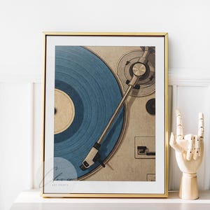 May include: A framed print featuring a close-up of a vintage record player with a blue vinyl record. The artwork is in a gold frame and is displayed on a white surface. A wooden hand sculpture is next to the frame.
