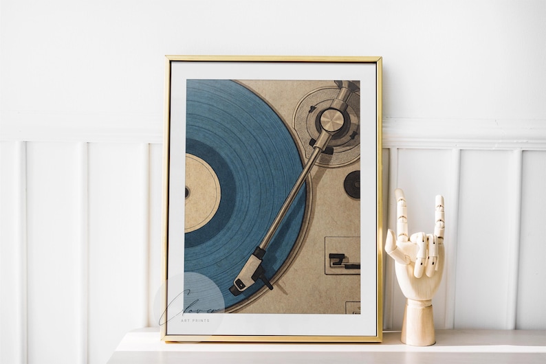 May include: A framed print featuring a close-up of a vintage record player with a blue vinyl record. The artwork is in a gold frame and is displayed on a white surface. A wooden hand sculpture is next to the frame.