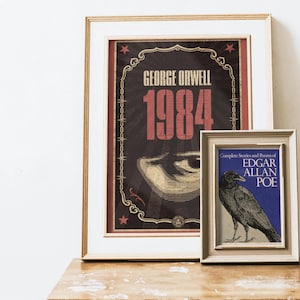 May include: Two framed prints on a wooden surface. The larger print features the text "GEORGE ORWELL 1984" in red and gold against a dark background. The smaller print displays a raven illustration and the text "EDGAR ALLAN POE".