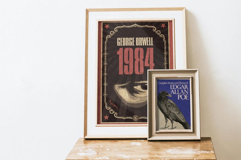 May include: Two framed prints on a wooden surface. The larger print features the text "GEORGE ORWELL 1984" in red and gold against a dark background. The smaller print displays a raven illustration and the text "EDGAR ALLAN POE".