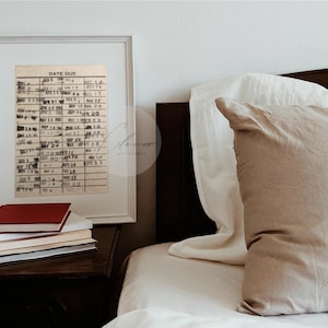 May include: A framed print of a vintage library card with the text "Date Due" and a grid of dates. The print is hanging on a white wall above a nightstand with a stack of books.