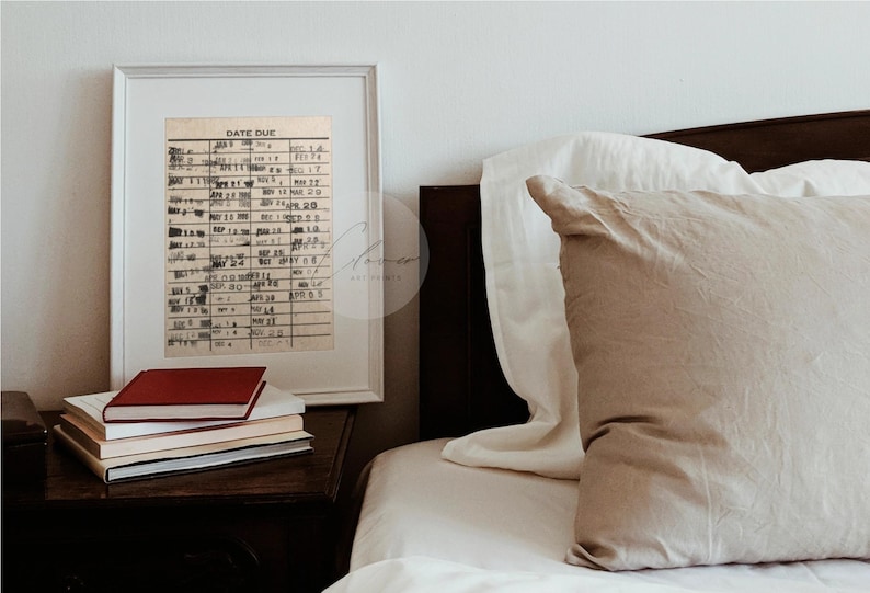 May include: A framed print of a vintage library card with the text "Date Due" and a grid of dates. The print is hanging on a white wall above a nightstand with a stack of books.