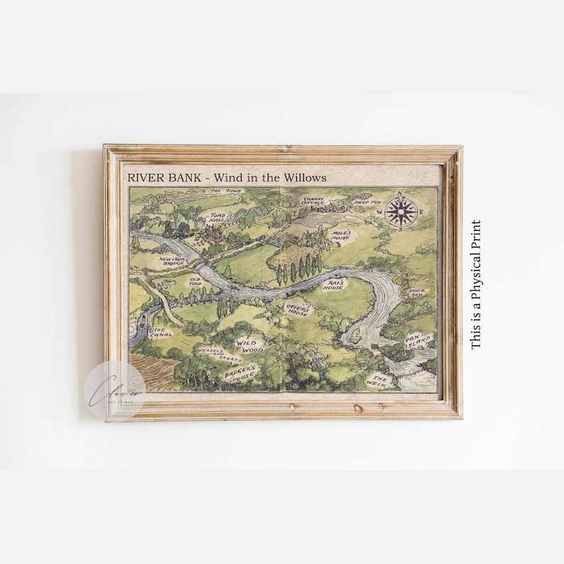 May include: A framed print of a map titled "River Bank - Wind in the Willows." The map features a river winding through a green landscape with labeled locations. A compass rose is in the upper right corner. The frame is a light brown color.