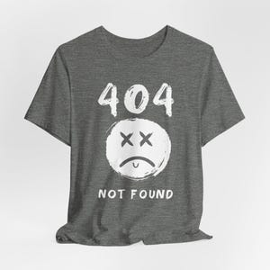 Könnte beinhalten: Dunkelgraues T-Shirt mit der Nummer "404" über einer weißen, im Used-Look gehaltenen traurigen Gesichts-Grafik. Unter der Grafik steht der Text "NOT FOUND" in Weiß. Das Shirt hat einen Rundhalsausschnitt und kurze Ärmel.