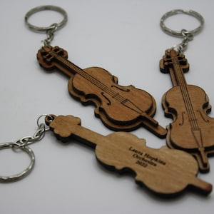 May include: Three wooden cello keychain ornaments with silver key rings. The keychains are engraved with the text "Laura Hopkins Orchestra 2022".
