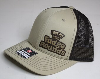 May include: A beige and black trucker hat with a brown patch that says "I buy my own Fudge Rounds".