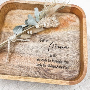 Può includere: Un vassoio quadrato in legno con finitura naturale, con la scritta "Danke Mama" e altro testo in tedesco. Il vassoio è decorato con un piccolo mazzo di fiori secchi e verde.