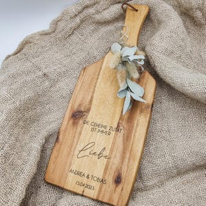 Può includere: Un tagliere di legno con manico, inciso con il testo "Die geheime Zutat ist immer Liebe Andrea & Tobias 15.04.2023". Il tagliere è decorato con un rametto di eucalipto essiccato.