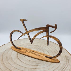 Op de afbeelding: Een bruine houten sculptuur van een fietssilhouet met een zichtbare houtnerf. De fiets is gemonteerd op een bijpassende ovale houten voet met een gegraveerde hartslaglijn die een klein fietspictogram en een hartsymbool bevat.