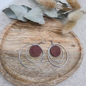 May include: A pair of earrings with a modern design. Each earring features a silver hoop with a smaller, inner hoop and a round, dark brown wooden disc. The earrings are displayed on a wooden tray.
