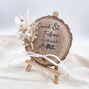 Könnte beinhalten: Eine runde Holzscheibe mit der Gravur Sarah & Fabian, dem Datum 13.06.2026 und zwei Vögeln auf einem Ast. Sie ist mit einem kleinen Trockenblumenstrauß und zwei goldenen Ringen an einem weißen Band verziert und steht auf einer kleinen Holzstaffelei.