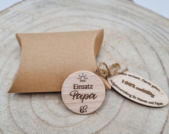 Könnte beinhalten: Ein Holz-Kreis mit dem Text "Einsatz Papa" und einem kleinen Symbol für einen Schnuller. Der Kreis ist an einer braunen Papiertüte mit einem Etikett befestigt, auf dem "100% wichtig" und "Aufgabenverteilung für Mamas und Papas" steht.