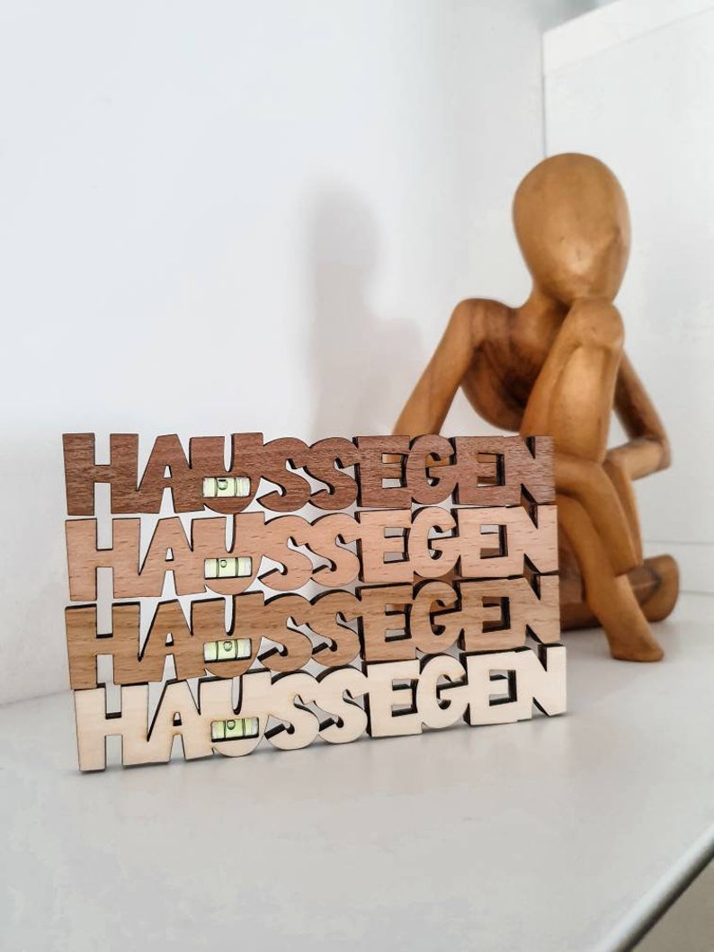 May include: Four wooden blocks stacked on top of each other, each block spelling out the word "Haus Segen" in a different shade of brown. The blocks are sitting on a white surface in front of a wooden figurine.