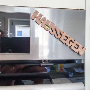 May include: A wooden sign with the word "Haussegen" in German, meaning "blessing of the house", is attached to a stainless steel oven door.