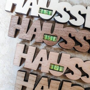 May include: Four wooden signs spelling "HAUS" with a small green level attached to each sign. The signs are stacked on top of each other, with the top sign being white, the second sign being brown, the third sign being light brown, and the bottom sign being a darker brown.