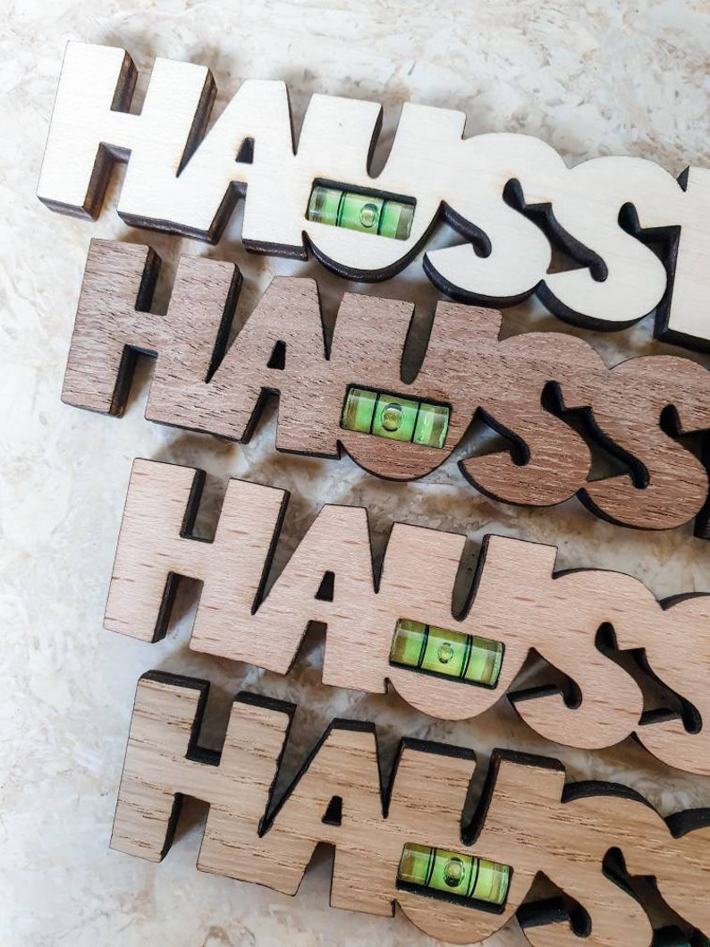 May include: Four wooden signs spelling "HAUS" with a small green level attached to each sign. The signs are stacked on top of each other, with the top sign being white, the second sign being brown, the third sign being light brown, and the bottom sign being a darker brown.