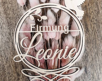 Pode incluir: Um topo de bolo de madeira com as palavras "Firmung Leonie" em uma fonte decorativa, com uma pomba e detalhes florais. O topo é colocado em um fundo de flores secas rosa, criando uma estética delicada e festiva.