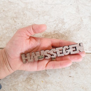 May include: A wooden sign with the word "HAUSSEGEN" carved into it. The sign is held in a hand.