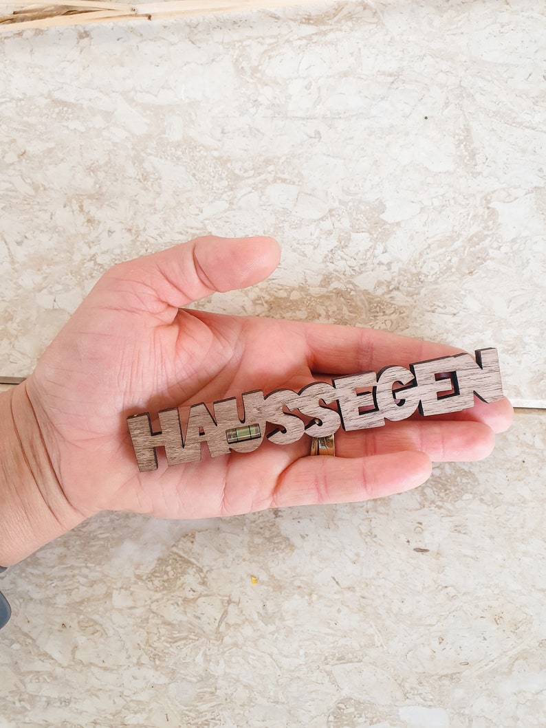 May include: A wooden sign with the word "HAUSSEGEN" carved into it. The sign is held in a hand.
