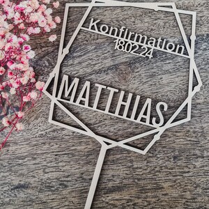 Op de afbeelding: Houten taarttopper met de tekst "Konfirmation 18.02.24" en de naam "MATTHIAS". De topper heeft een geometrisch ontwerp en een vissymbool. Roze bloemen staan op de achtergrond.
