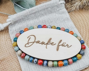 Puede incluir: Un letrero ovalado de madera con el texto "Danke für" rodeado por un borde de cuentas de colores. El letrero está sobre una bolsa de tela marrón claro.
