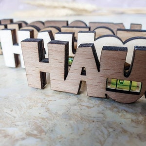 May include: A close-up of a wooden sign spelling "HAUS" with a small level embedded in the "U". The sign is made of light brown wood with dark brown edges.